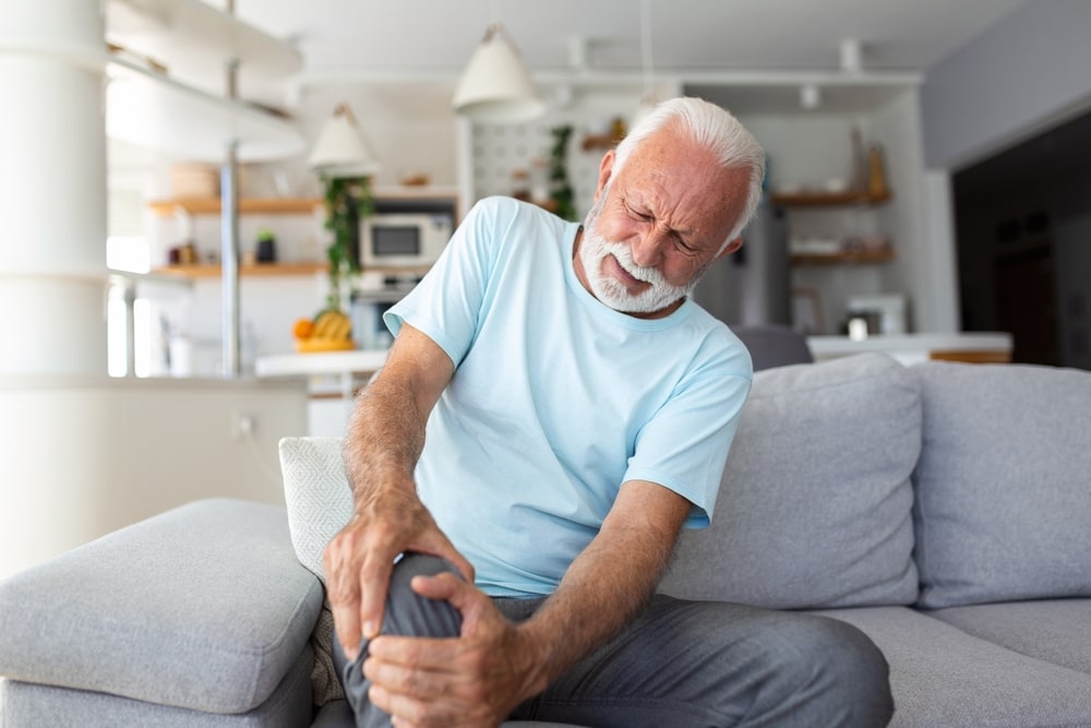 The older man is sitting on the couch has pain in the knee joint